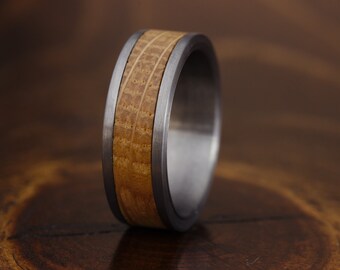 Puede incluir: Un anillo que presenta una banda de metal gris cepillado con una incrustación central de madera de color marrón claro. La incrustación de madera muestra un patrón de veta distintivo y está flanqueada por dos bordes metálicos finos.