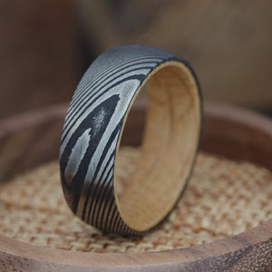 Op de afbeelding: Een metalen ring met een zwart en zilveren patroon en een houten band. De ring ligt op een houten oppervlak.