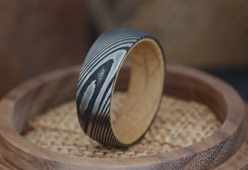 Op de afbeelding: Een metalen ring met een zwart en zilveren patroon en een houten band. De ring ligt op een houten oppervlak.