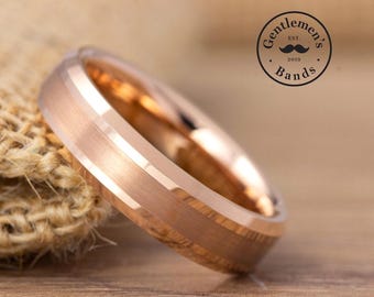 May include: A rose gold-colored ring with a brushed finish and beveled edges. The ring features a circular band design. In the upper right corner, a circular logo contains the text "Gentlemen's Bands" with an illustration of a mustache.