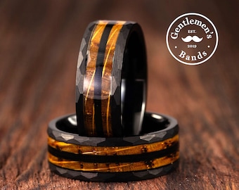 May include: Two black rings with a hammered finish and a central inlay of brown wood grain. The rings feature a dark, textured exterior and a smooth, polished interior. A circular logo in the top right corner reads Gentlemen's Bands Est. 2019.