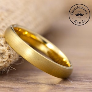 May include: A gold-colored ring with a brushed matte finish on the outer surface and a polished, reflective interior. The ring is positioned near a textured fabric. A circular logo in the upper right corner reads Gentleman's Bands EST. 2019.