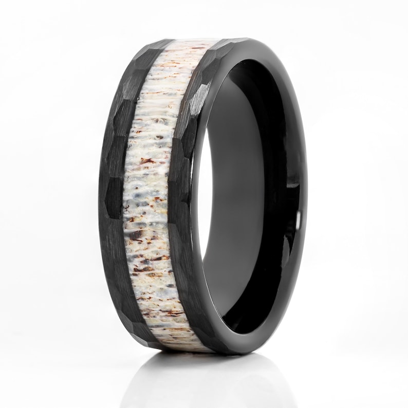 May include: A black ring featuring a hammered finish on the outer edges and a central inlay of textured, cream-colored organic material with brown and tan flecks. The band has a smooth, polished black interior surface and a wide, flat profile.