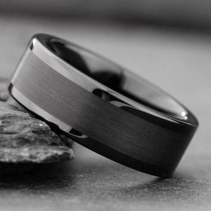 May include: A black ring with a brushed center band and polished edges. The ring is made of a dark material and is set against a gray background. The ring is a wide band style.
