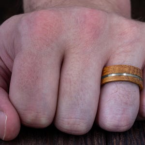 May include: A ring featuring a band made of light brown wood with a visible grain pattern, bisected horizontally by a thin, smooth, metallic silver-colored inlay that runs around the entire circumference of the circular jewelry piece.
