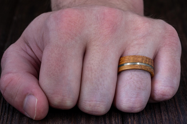 May include: A ring featuring a band made of light brown wood with a visible grain pattern, bisected horizontally by a thin, smooth, metallic silver-colored inlay that runs around the entire circumference of the circular jewelry piece.