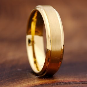 May include: A gold-colored ring featuring a wide, brushed matte center band flanked by two polished, reflective beveled edges. The band has a consistent width and a smooth, metallic finish that catches the light along its outer rim.