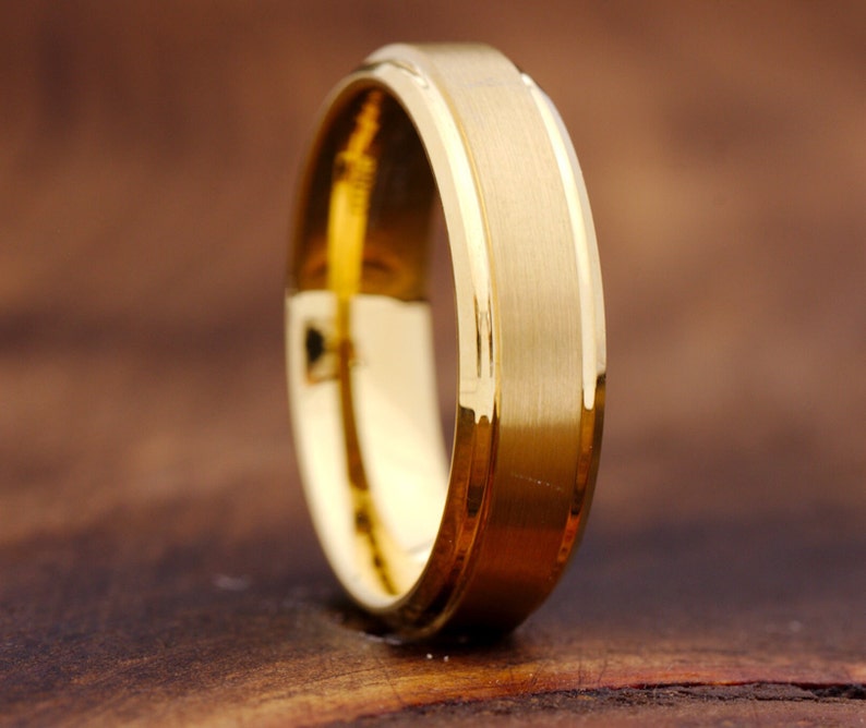 May include: A gold-colored ring featuring a wide, brushed matte center band flanked by two polished, reflective beveled edges. The band has a consistent width and a smooth, metallic finish that catches the light along its outer rim.