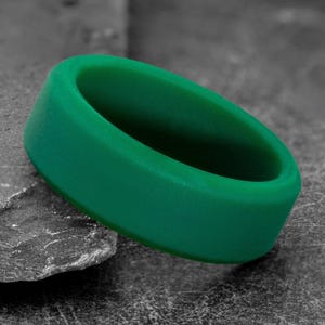 May include: A vibrant green silicone ring. The ring is a solid, smooth band with a rounded interior and a flat exterior. The ring is resting on a gray, textured surface.