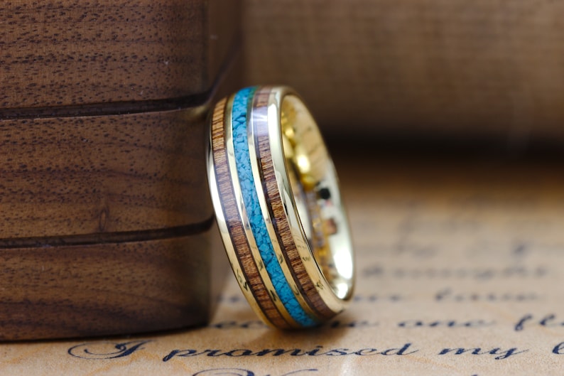 May include: A gold ring with a band of wood and turquoise inlay. The ring is on a piece of paper with the text "I promised my l" visible.