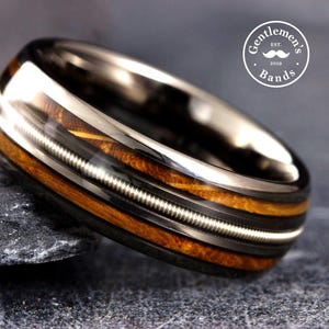 May include: A polished dark metal ring featuring a central inlay of brown wood grain and a thin silver-colored guitar string wrapped around the circumference. The ring displays a circular logo in the upper right corner with the text Gentlemen's Bands Est. 2019.