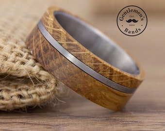 Puede incluir: Un anillo con una banda exterior de madera marrón texturizada y una incrustación fina de metal plateado en el centro. El interior del anillo es de metal plateado liso. Un logotipo circular muestra el texto Gentleman's Bands est. 2019.