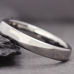 Può includere: Un anello argentato lucido con una superficie liscia e arrotondata. L'anello è posto su uno sfondo grigio scuro, evidenziando le sue qualità riflettenti. Il design dell'anello è semplice ed elegante, adatto all'uso quotidiano.