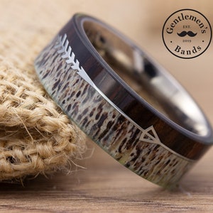 Puede incluir: Un anillo con una incrustación de madera oscura y una incrustación de asta texturizada de color claro, acentuado por un diseño de flecha en tono plateado. La banda tiene un interior liso y pulido. Un logo circular con el texto Gentleman's Bands Est. 2019.