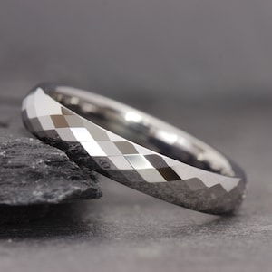 May include: A silver tungsten carbide ring with a faceted, diamond-cut design. The ring is on a grey background.