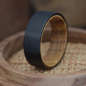 Op de afbeelding: Een zwarte ring van wolfraamcarbide met een houten inlay. De ring ligt op een houten oppervlak.