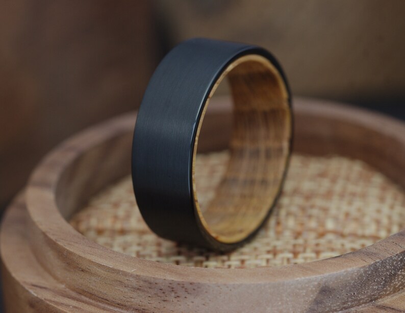Op de afbeelding: Een zwarte ring van wolfraamcarbide met een houten inlay. De ring ligt op een houten oppervlak.