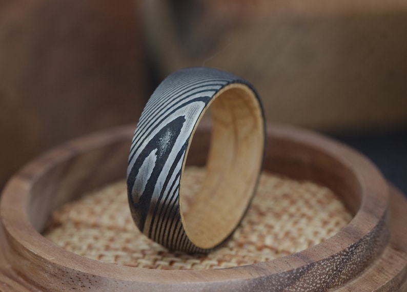 May include: A black and silver patterned ring with a wooden band. The ring is sitting on a wooden surface.