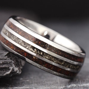 May include: A silver-toned metal ring featuring three horizontal channels. The outer two channels are filled with a dark reddish-brown granular material, while the center channel contains a shimmering, metallic grey textured inlay.