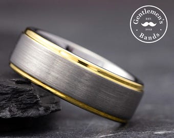 May include: A close-up of a tungsten ring with a brushed silver finish and a gold-colored edge. The ring is set against a dark background. The ring has the text "Gentlemen's Bands" and the year "2019" in a circular logo.