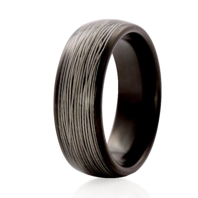 Op de afbeelding: Een zwarte ring met een uniek ontwerp. De ring heeft een donkere band met een patroon van dunne, parallelle lijnen in een lichtere kleur, waardoor een textuur ontstaat. De ring staat tegen een witte achtergrond.