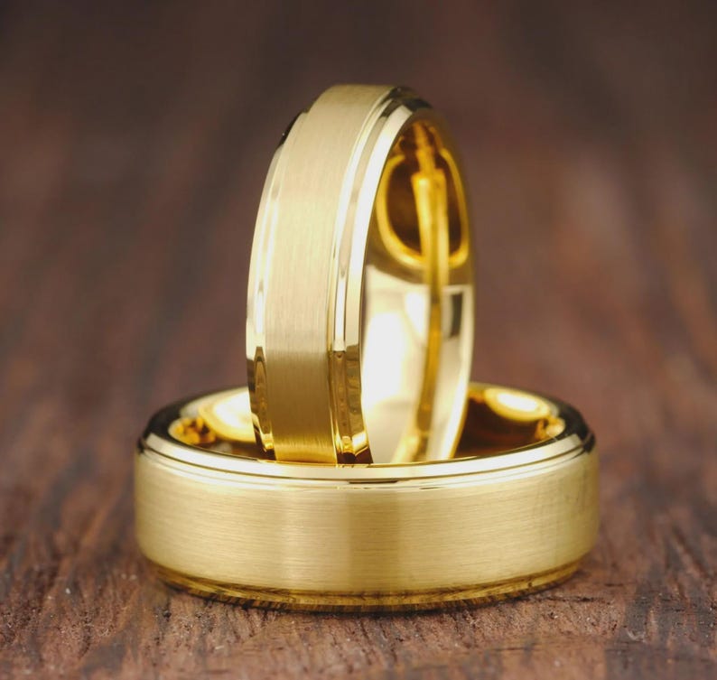 May include: Two gold-colored rings with a brushed matte finish on the outer surface and polished, shiny edges. One ring is positioned vertically, resting inside the center of the other ring, which lies flat in a horizontal orientation.