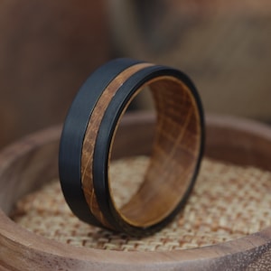 May include: A black tungsten carbide ring with a brown wood inlay. The ring is on a wooden display stand.