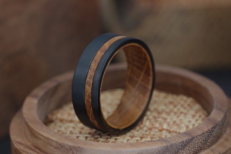 May include: A black tungsten carbide ring with a brown wood inlay. The ring is on a wooden display stand.