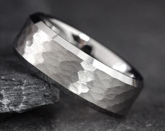 May include: A silver-colored ring with a hammered texture on the band. The ring has a polished, beveled edge and a smooth, reflective interior. The ring is set against a dark background.