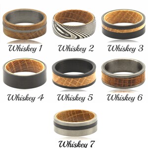 May include: Seven different styles of men's rings featuring a whiskey barrel wood inlay. The rings are crafted from various metals, including stainless steel, black titanium, and Damascus steel. The wood inlay is a light brown colour and displays a natural grain pattern. The rings are all of varying sizes and widths.