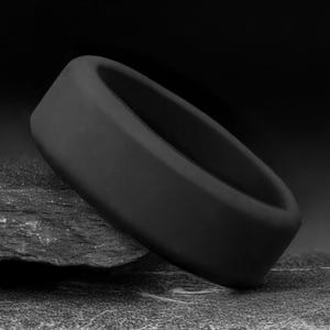 Op de afbeelding: Een effen zwarte ring met een gladde, matte afwerking en een afgerond profiel. De band heeft een dikke, uniforme breedte en een eenvoudig, onversierd ontwerp. De ring staat in een hoek, waardoor de cirkelvorm en donkere kleur zichtbaar zijn.