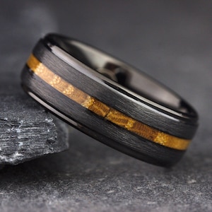 May include: A black ring with a brushed finish featuring a central inlay of textured, golden-brown fossilized material. The band has a smooth, polished interior and a dark, matte exterior surface that contrasts with the organic, granular pattern of the inlay.