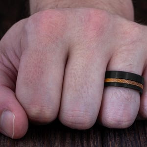 May include: A matte black ring featuring a central horizontal inlay of light brown wood grain. The band has a flat profile and a smooth finish, encircling the finger with a distinct contrast between the dark metal and the natural wood texture.
