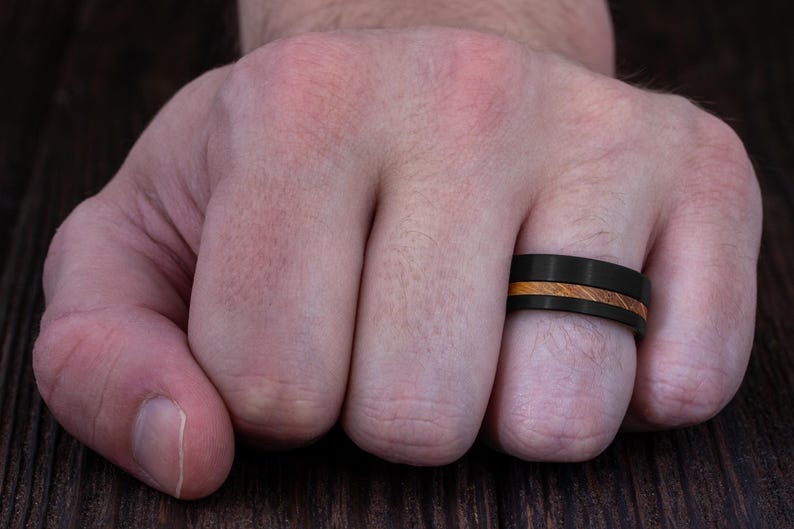 May include: A matte black ring featuring a central horizontal inlay of light brown wood grain. The band has a flat profile and a smooth finish, encircling the finger with a distinct contrast between the dark metal and the natural wood texture.
