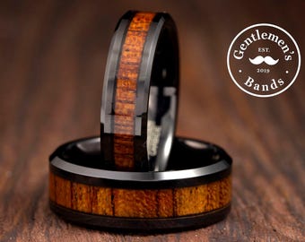 May include: Two rings with a black metal finish and a central inlay of brown wood grain. One ring stands vertically while the other lies flat. A circular logo in the top right corner features the text "Gentlemen's Bands EST. 2019" with a mustache icon.