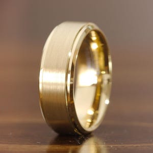 May include: A gold-colored ring with a brushed matte finish on the outer band and polished, beveled edges. The band has a flat profile and a reflective interior surface, showcasing a metallic luster and a clean, simple design throughout the piece.