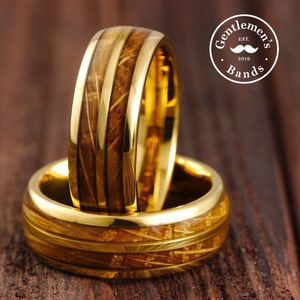 May include: Two gold wedding bands with a wood inlay design. The bands are stacked on top of each other. The top band has a wider wood inlay than the bottom band. The bands are on a wooden surface. The text "Gentlemen's Bands Est. 2019" is visible in the background.