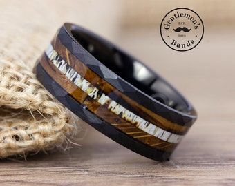 May include: A black faceted ring featuring a central inlay of brown wood and white antler material. The band has a textured, geometric surface. A circular logo in the upper right corner displays the text Gentlemen's Bands Est. 2019 with a mustache.