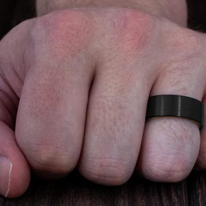 May include: A wide, matte black band ring with a smooth, brushed finish. The ring features a flat profile and a solid, dark metallic appearance, encircling the finger with a consistent width and a clean, unadorned surface design.
