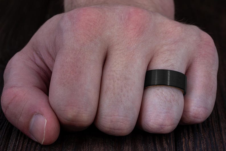 May include: A wide, matte black band ring with a smooth, brushed finish. The ring features a flat profile and a solid, dark metallic appearance, encircling the finger with a consistent width and a clean, unadorned surface design.