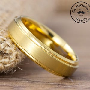 May include: A gold-colored ring with a brushed matte finish and polished beveled edges. The band features a central flat surface with two distinct grooves running along the circumference. A circular logo with the text Gentleman's Bands Est. 2019 is visible.