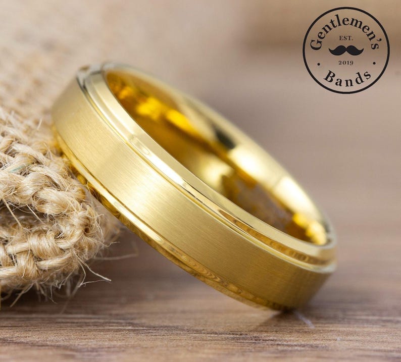 May include: A gold-colored ring with a brushed matte finish and polished beveled edges. The band features a central flat surface with two distinct grooves running along the circumference. A circular logo with the text Gentleman's Bands Est. 2019 is visible.