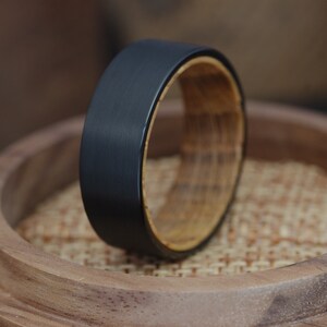 May include: A black tungsten carbide ring with a wooden inlay. The ring is sitting on a wooden surface.
