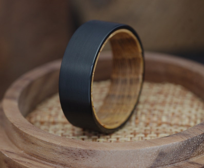 May include: A black tungsten carbide ring with a wooden inlay. The ring is sitting on a wooden surface.