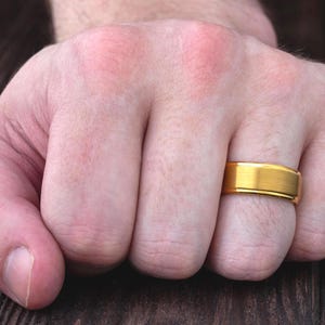 May include: A gold-colored ring with a brushed matte finish on the center band and polished, shiny edges. The ring features a flat profile and a simple, clean design, worn on the ring finger of a hand positioned against a dark wooden surface.