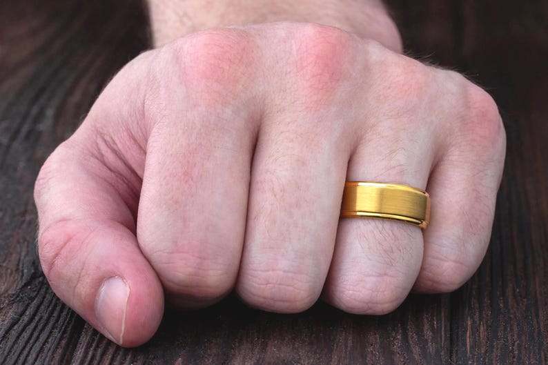 May include: A gold-colored ring with a brushed matte finish on the center band and polished, shiny edges. The ring features a flat profile and a simple, clean design, worn on the ring finger of a hand positioned against a dark wooden surface.
