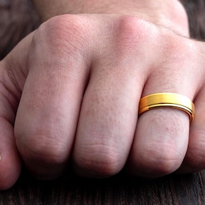 May include: A gold-colored ring with a smooth, brushed finish and a flat profile. The band features a subtle central groove that runs horizontally around the circumference, creating a clean and simple design for this metallic finger accessory.