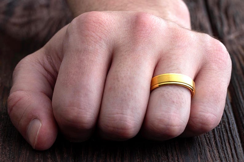 May include: A gold-colored ring with a smooth, brushed finish and a flat profile. The band features a subtle central groove that runs horizontally around the circumference, creating a clean and simple design for this metallic finger accessory.