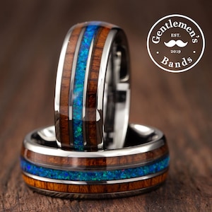 May include: Two rings featuring a polished silver-tone metal band with a central inlay of vibrant blue crushed opal flanked by two strips of dark brown wood grain. A circular logo in the upper right corner reads Gentlemen's Bands EST. 2019.
