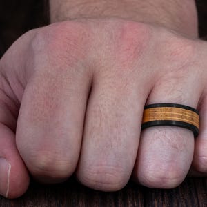 May include: A ring featuring a matte black band with a central inlay of light brown wood. The wood inlay is divided into three horizontal sections by two thin black lines, creating a layered appearance around the circumference of the ring.
